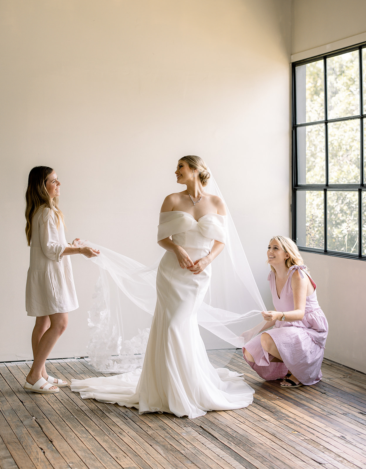 Two women help the bride get ready near to the window