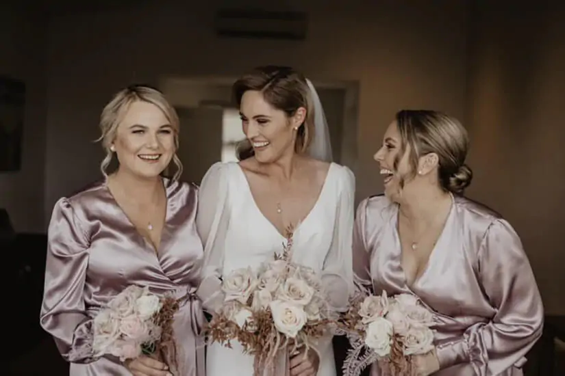 Happy bride wears white dress and bridesmaid wears light pink with flowers